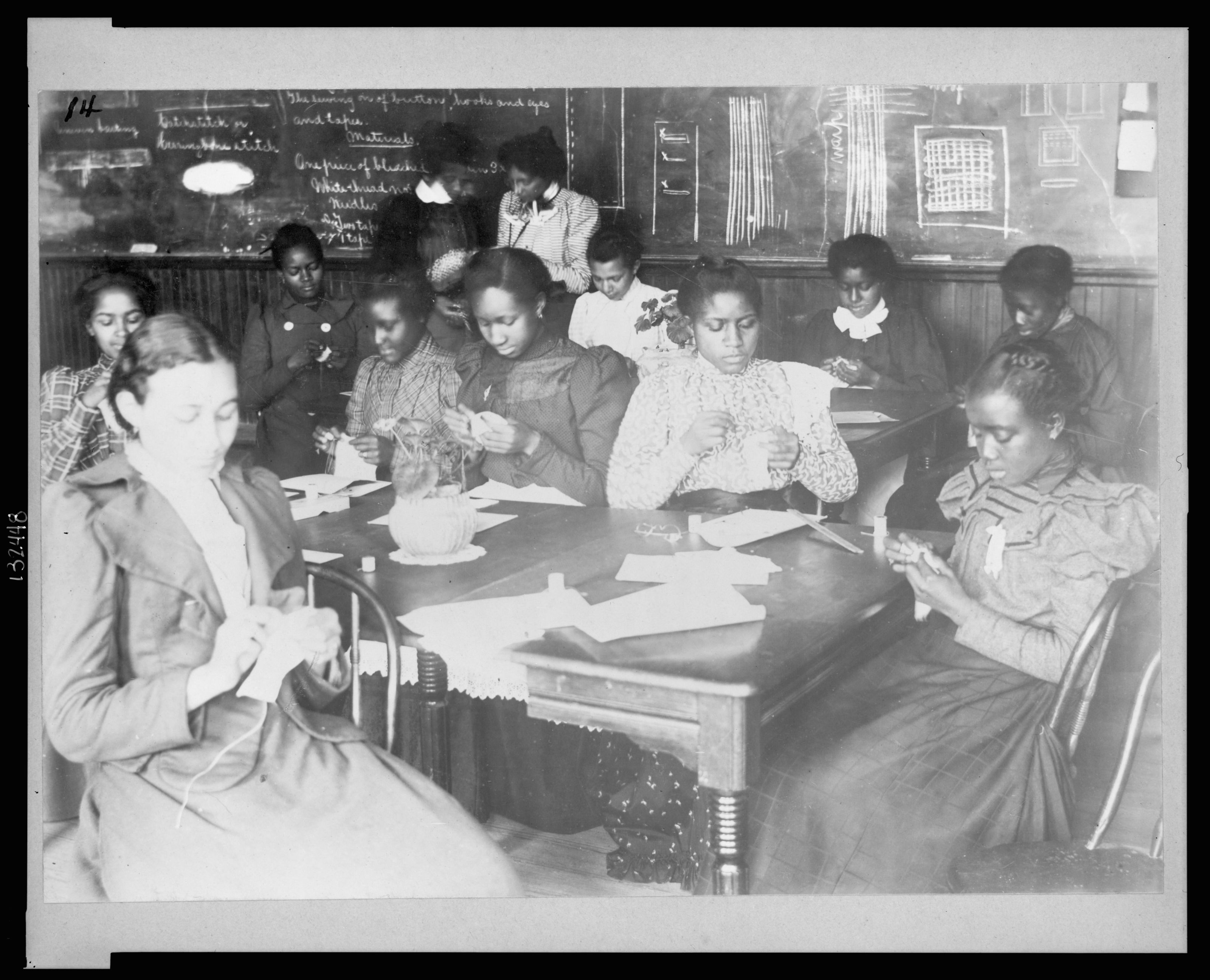 A black and white photograph of a classroom of Black female students practice sewing by hand. They wear long dresses and are seated around a wood table.